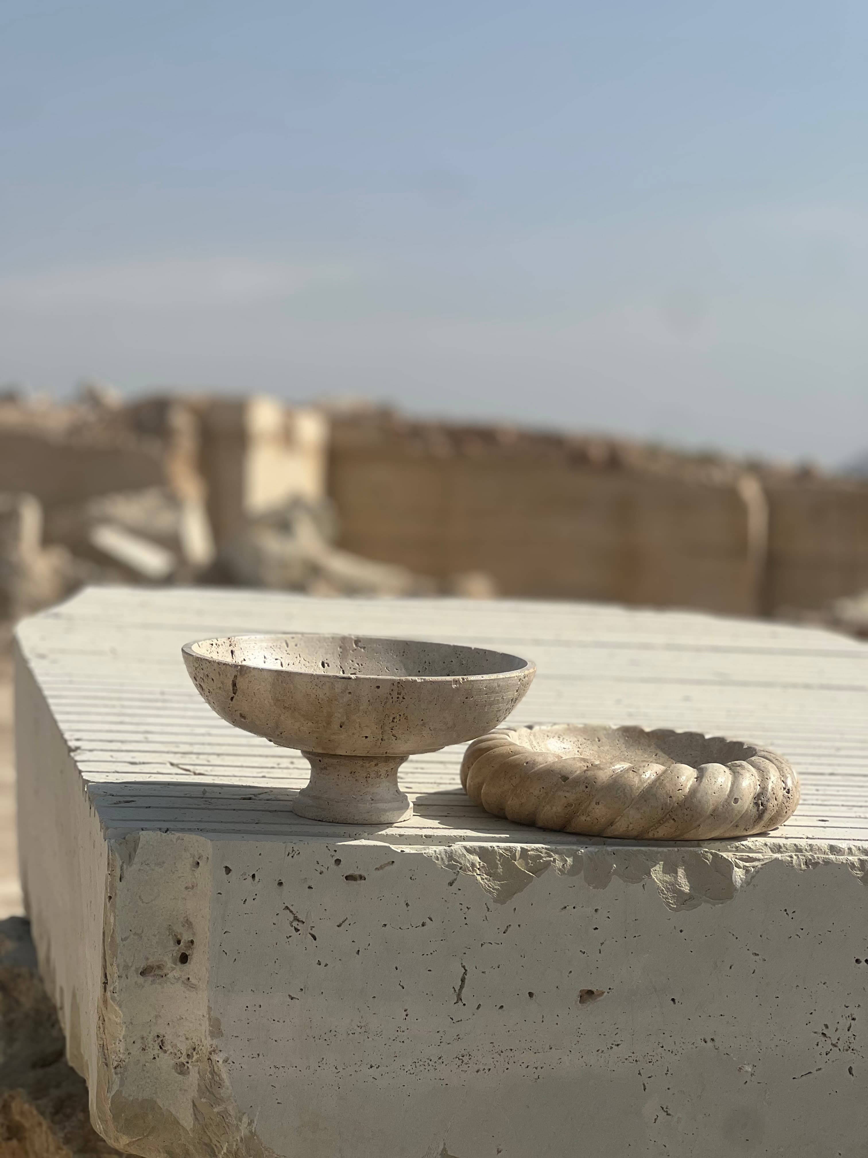 Two hand-finished travertine bowls resting on a raw quarry block, with cut blocks in the background
